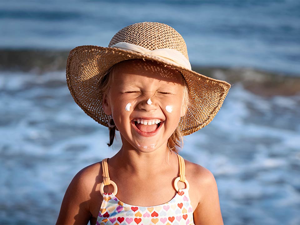 Lachendes Kind steht am Meer und hat Sonnencreme im Gesicht.