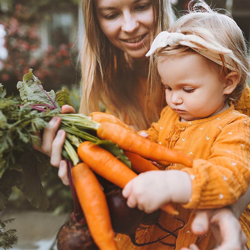 Eine Mutter sitzt mit ihrer Tochter im Garten und hält einen Bund Karotten in der Hand.