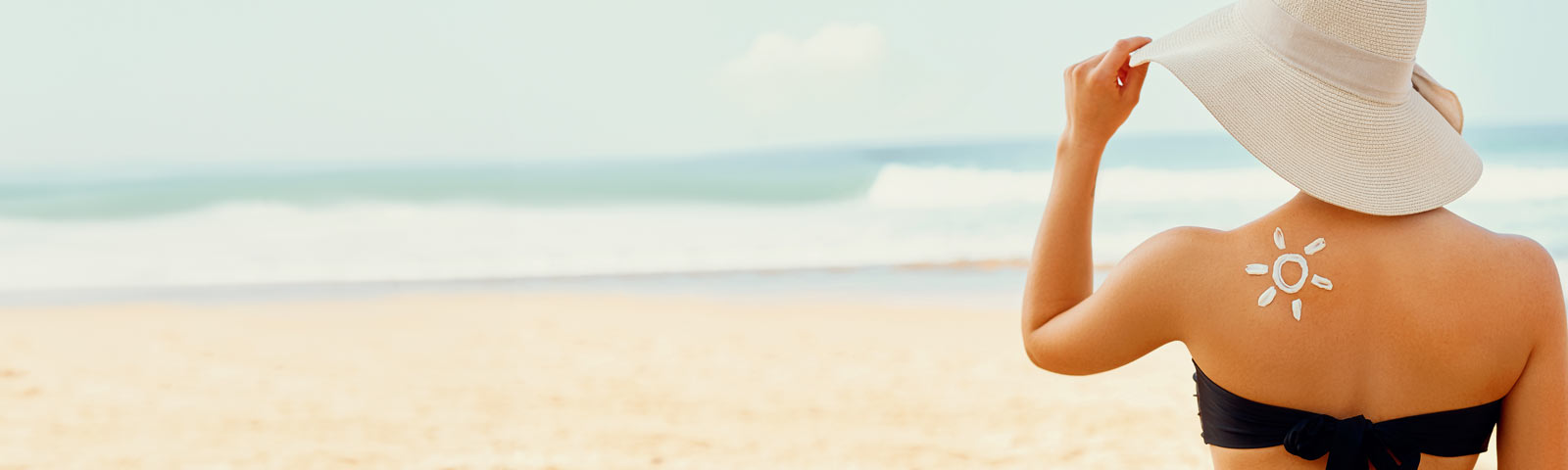 Eine Frau steht am Strand, genießt die Sonne und trägt zum Schutz vor der Sonnen einen großen Hut.