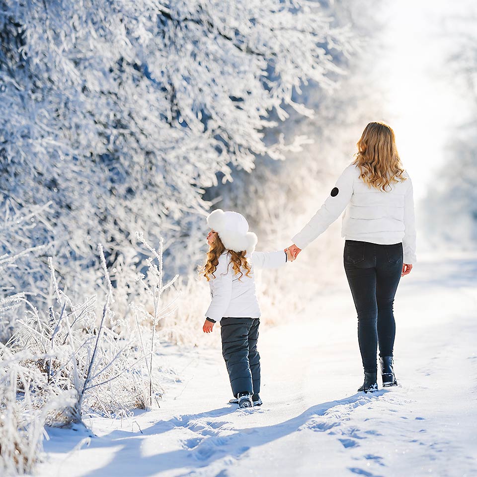 Eine Frau spaziert mit ihrer Tochter durch eine Schneelandschaft am Wald mit Sonnenschein.
